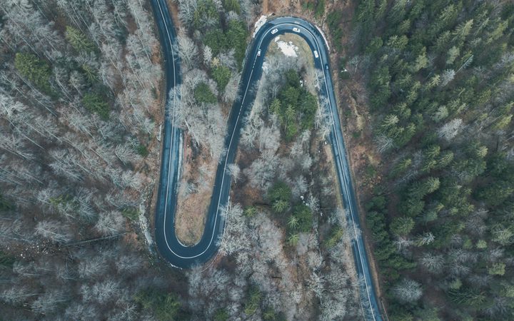 A winding mountain road through a dense forest, with some trees showing winter leaflessness, captured from an aerial perspective.