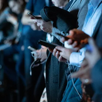 People in business attire are focused on their smartphones in a dark, indoor setting, likely at a conference or event.