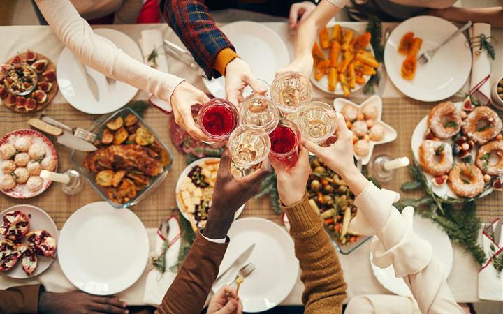 Top-down view of a diverse group toasting with wine glasses around a festive table filled with snacks, pastries and roasted dishes.