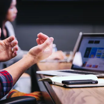 Hands gesturing in discussion, with a laptop and notebook on a wooden table during a meeting or presentation.