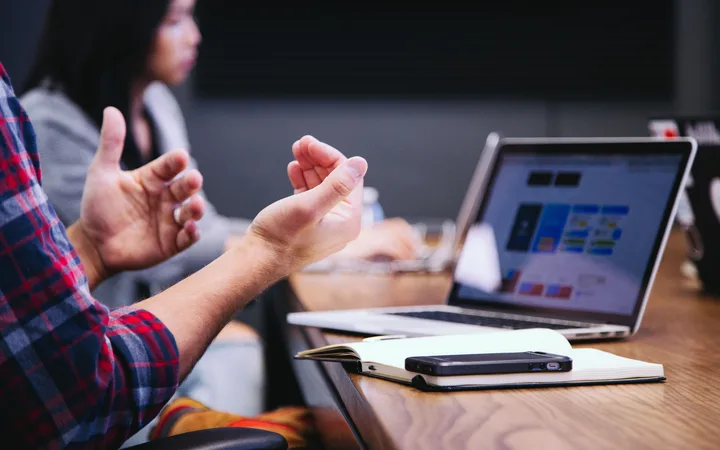 Hands gesturing in discussion, with a laptop and notebook on a wooden table during a meeting or presentation.