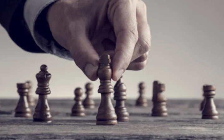 A hand moving a chess piece on a chessboard, with several other pieces visible in the background.