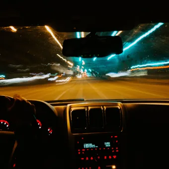 Inside a moving car at night, view of the dashboard and blurred city lights through the windshield with streaks of blue, orange, and white lights.