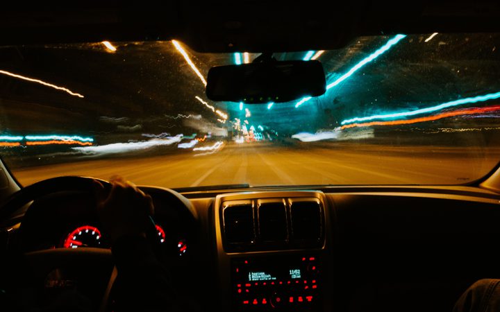Inside a moving car at night, view of the dashboard and blurred city lights through the windshield with streaks of blue, orange, and white lights.