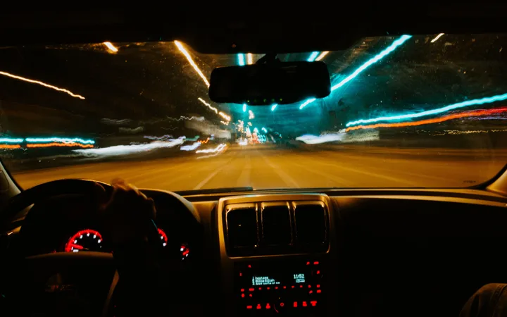 Inside a moving car at night, view of the dashboard and blurred city lights through the windshield with streaks of blue, orange, and white lights.