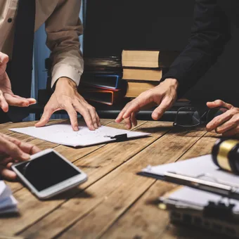 People collaborating around a wooden table with papers, books, a smartphone, and headphones, engaging in a discussion or brainstorming session.