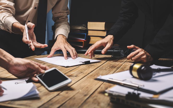 Hands of multiple people discussing and working on documents and papers at a wooden table, with books, a phone, and headset around.
