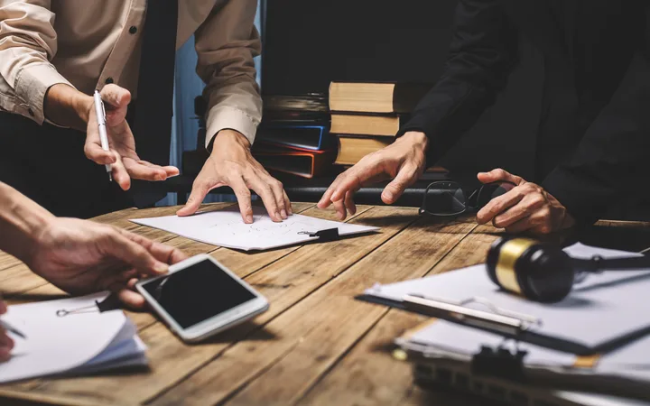 Hands of multiple people discussing and working on documents and papers at a wooden table, with books, a phone, and headset around.