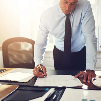 A person in a white shirt and black tie leans over a desk, writing on documents with a coffee cup nearby in a bright office setting.