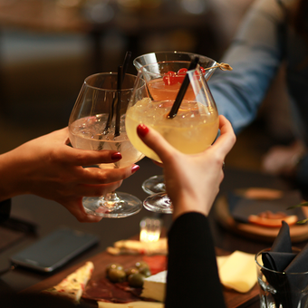 People clink stemmed cocktails over a cheese board with olives and a candlelit table in a dim restaurant.