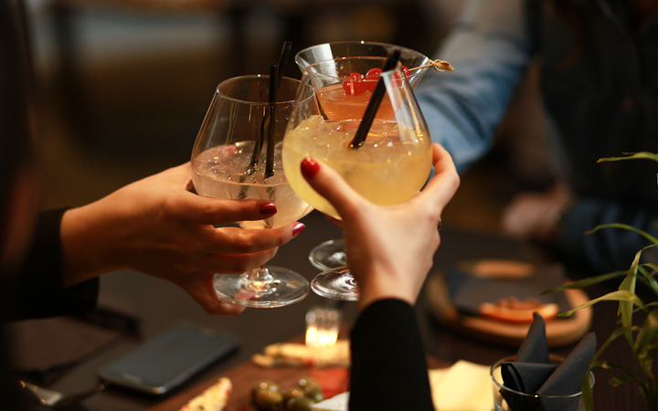 People clink stemmed cocktails over a cheese board with olives and a candlelit table in a dim restaurant.