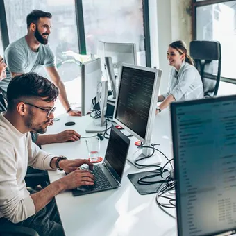 Bright open-plan office with several people at desks; a person stands and chats while others code on multiple monitors.