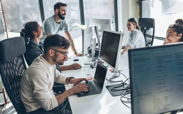 Five colleagues in a bright open-plan office at shared desks with multiple monitors; one person stands and chats while others code.