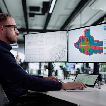 Portrait of a smiling person with short gray hair and glasses, wearing a red and black patterned top, against a gray backdrop.