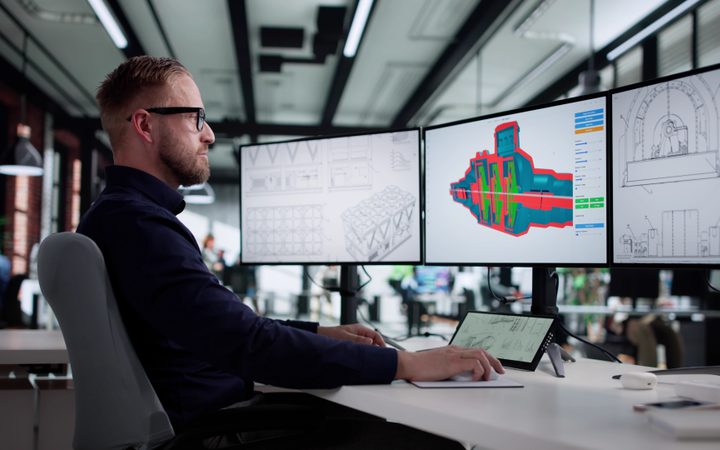 Side profile of a person with glasses and a beard at a desk with 3 monitors showing technical drawings and a colourful 3D model in bright office.