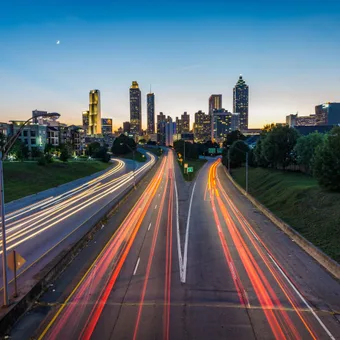 City skyline at dusk with light trails on busy roads heading toward the tall buildings and a clear sky with a crescent moon.