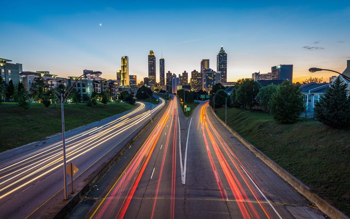 City skyline at dusk with light trails on busy roads heading toward the tall buildings and a clear sky with a crescent moon.