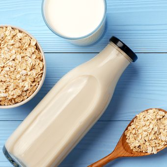 Flat lay of a bottle of oat milk, a glass of milk, a bowl of rolled oats and a wooden spoon with oats on a light blue wooden surface.