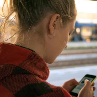 Young person in a red checkered hoodie looking at her phone, with a blurred train station platform and passing trains in the background.