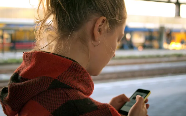 Young person in a red checkered hoodie looking at her phone, with a blurred train station platform and passing trains in the background.