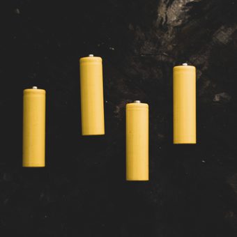 Four yellow candles are arranged on a dark, textured surface, with some candles standing upright and one slightly tilted.