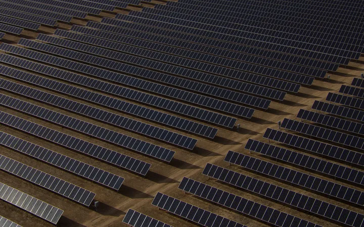 Aerial view of numerous dark solar panels arranged in parallel rows on a brown earth surface, creating a pattern across the landscape.