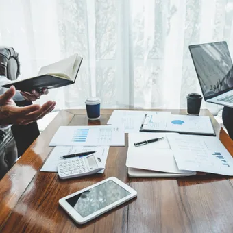 Two people in business attire sitting at a wooden table with documents, a calculator, a tablet, and coffee cups during a meeting.