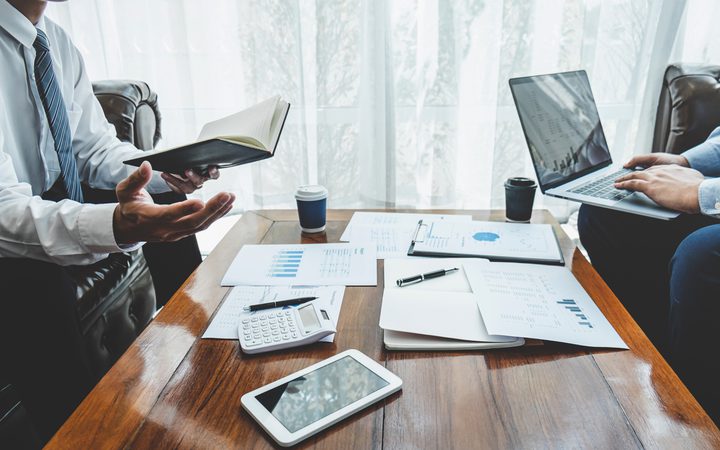 Two individuals in business attire are engaged in a meeting, with documents, a calculator, tablet, and laptops on a wooden table in a bright office.