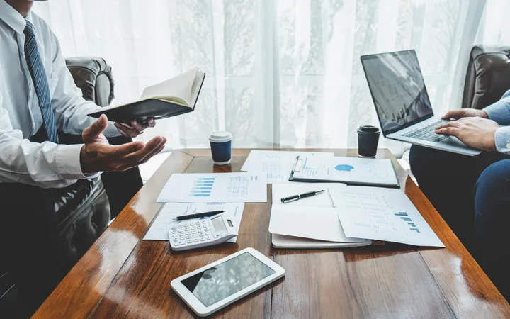 Two people in business attire sitting at a wooden table with documents, a calculator, a tablet, and coffee cups during a meeting.