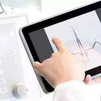 A healthcare professional interacts with a tablet displaying an electrocardiogram while monitoring medical equipment in a clinical setting.