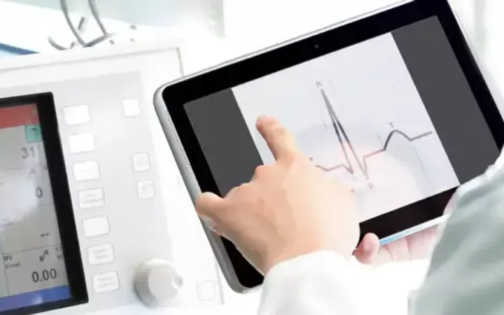 A healthcare professional interacts with a tablet displaying an electrocardiogram while monitoring medical equipment in a clinical setting.