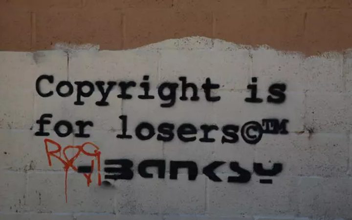 Graffiti on a wall reads "Copyright is for losers" with some graffiti tags, black stencil font on a brick wall background.