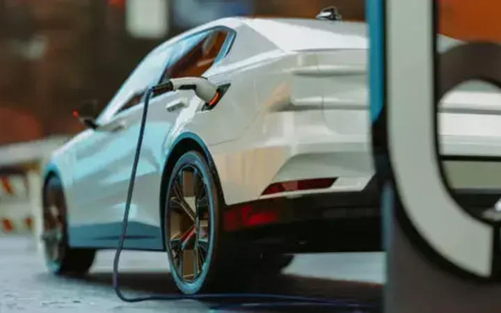 An electric car plugged into a charging station outdoors with a blurred city background.