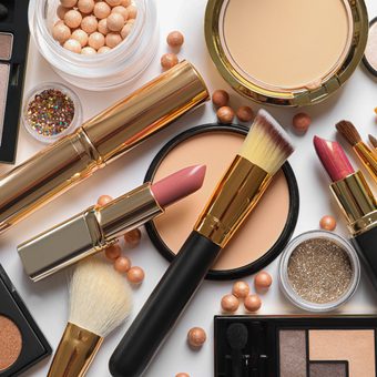 Assorted makeup items scattered on a white background: lipsticks, brushes, powder compacts, an eyeshadow palette, glitter, and small beads.