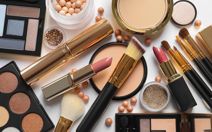 Assorted makeup items scattered on a white background: lipsticks, brushes, powder compacts, an eyeshadow palette, glitter, and small beads.