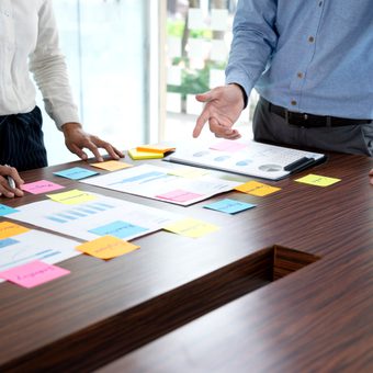 Two people stand around a wooden conference table, examining charts and post-it notes scattered across documents in a collaborative meeting.