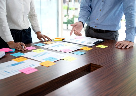 Deux personnes debout autour d'une table en bois, examinant des graphiques et documents, avec des post-its colorés éparpillés.