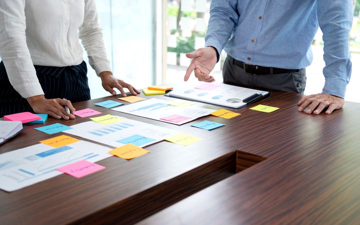 Two people stand around a wooden conference table, examining charts and post-it notes scattered across documents in a collaborative meeting.