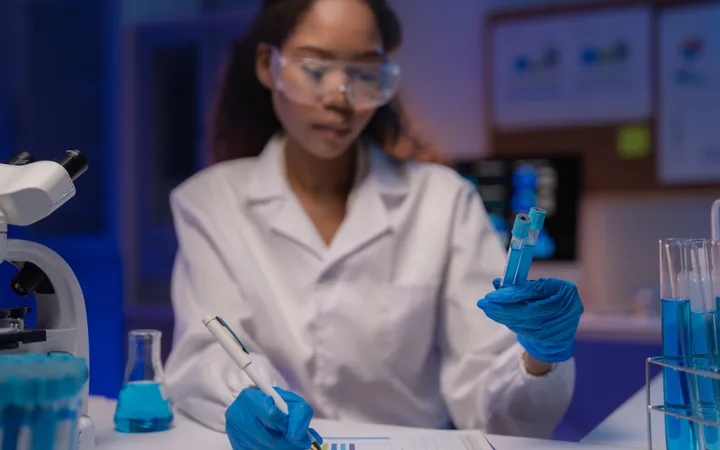 Scientist in a lab coat and safety goggles holding test tubes with blue liquid, surrounded by lab equipment in a modern scientific laboratory.