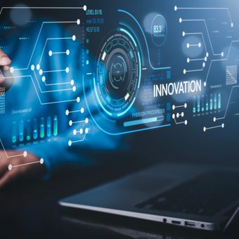 A hand interacts with a blue holographic interface showing gears, charts and the word INNOVATION, with a laptop in the background.