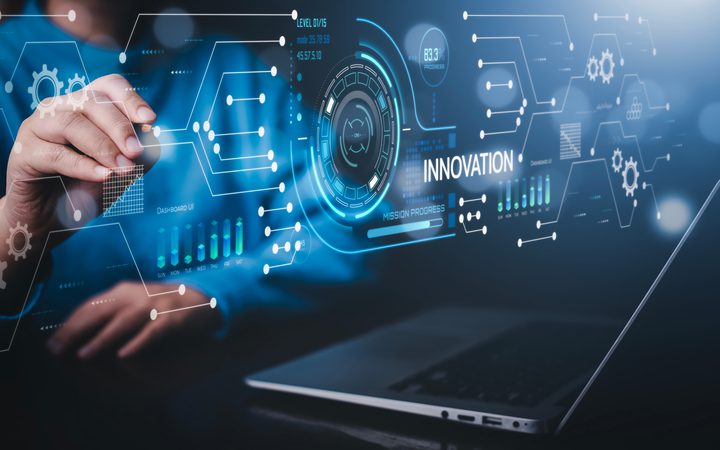 A hand interacts with a blue holographic interface showing gears, charts and the word INNOVATION, with a laptop in the background.