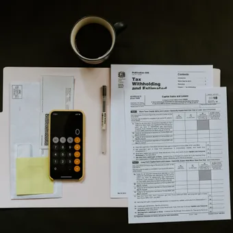 A desk with tax documents, a calculator on a notepad with sticky notes, a pen, a coffee mug, and a smartphone displaying a calculator app.