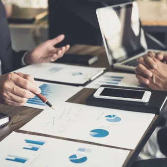 Two professionals discussing financial charts and data during a business meeting, with laptops, tablets, and printed graphs on the table.