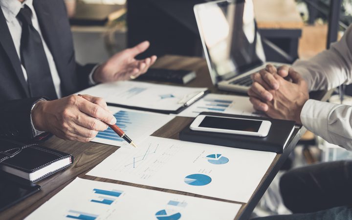 Two professionals discussing financial charts and data during a business meeting, with laptops, tablets, and printed graphs on the table.