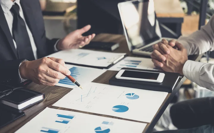Two professionals discussing financial charts and data during a business meeting, with laptops, tablets, and printed graphs on the table.