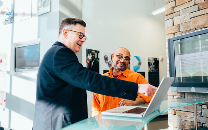 Two people are engaged in a conversation over a laptop in a modern office, smiling and gesturing, with a brick wall and monitors in the background.