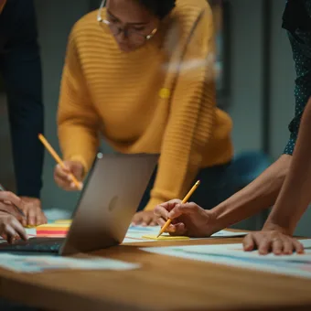 People gathered around a table working on papers with a laptop, holding pens and appearing focused on a collaborative task.