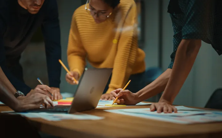 People gathered around a table working on documents, using laptops and holding pencils in a collaborative meeting or discussion.