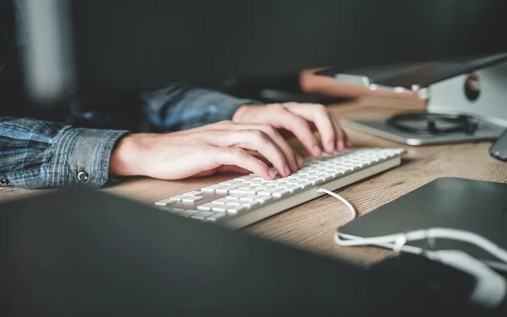Hands typing on a white keyboard at a wooden desk, with a monitor, mouse, and other desk items visible in the background.