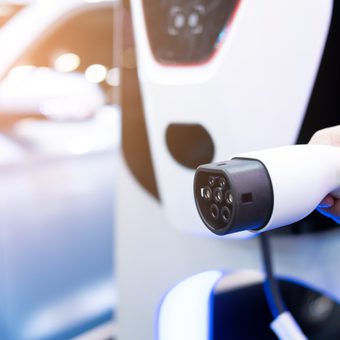 Close-up of a hand holding a white electric vehicle charging gun aimed at a car, with focus on the charging connector and port.